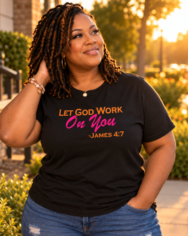 A woman wearing a black graphic t-shirt with the text 'LET GOD WORK On You -JAMES 4:7' in orange and pink lettering, paired with blue jeans, photographed outdoors in warm golden hour lighting.