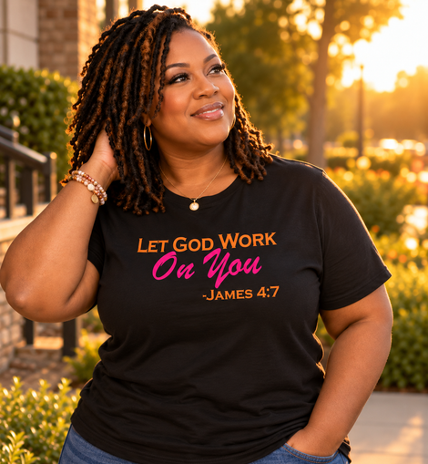 A woman wearing a black graphic t-shirt with the text 'LET GOD WORK On You -JAMES 4:7' in orange and pink lettering, paired with blue jeans, photographed outdoors in warm golden hour lighting.