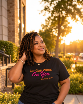 A woman wearing a black graphic t-shirt with the text 'LET GOD WORK On You -JAMES 4:7' in orange and pink lettering, paired with blue jeans, photographed outdoors in warm golden hour lighting.