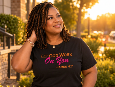A woman wearing a black graphic t-shirt with the text 'LET GOD WORK On You -JAMES 4:7' in orange and pink lettering, paired with blue jeans, photographed outdoors in warm golden hour lighting.