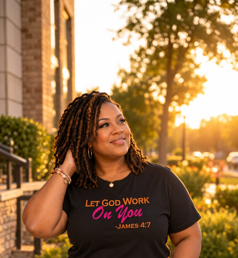 A woman wearing a black graphic t-shirt with the text 'LET GOD WORK On You -JAMES 4:7' in orange and pink lettering, paired with blue jeans, photographed outdoors in warm golden hour lighting.