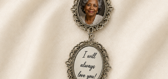 A silver-toned cremation keepsake brooch featuring a pearl-beaded safety pin, an oval photo frame with a portrait, an oval charm with the words 'I will always love you!', and a dangling angel figure, displayed on a cream satin fabric background.